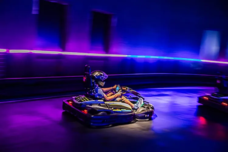 Person driving go-kart on indoor track with neon blue and purple lighting.