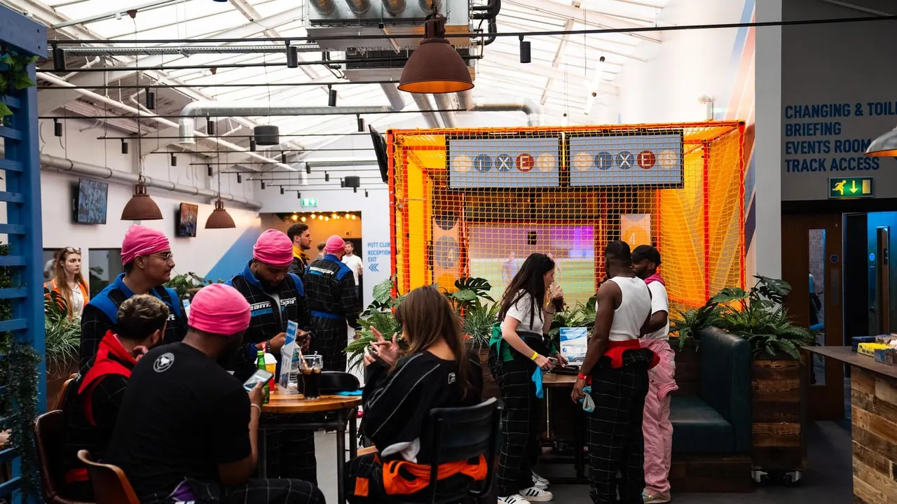 Groups of people socialising inside an indoor cricket venue with an orange-netted batting cage and bar area.