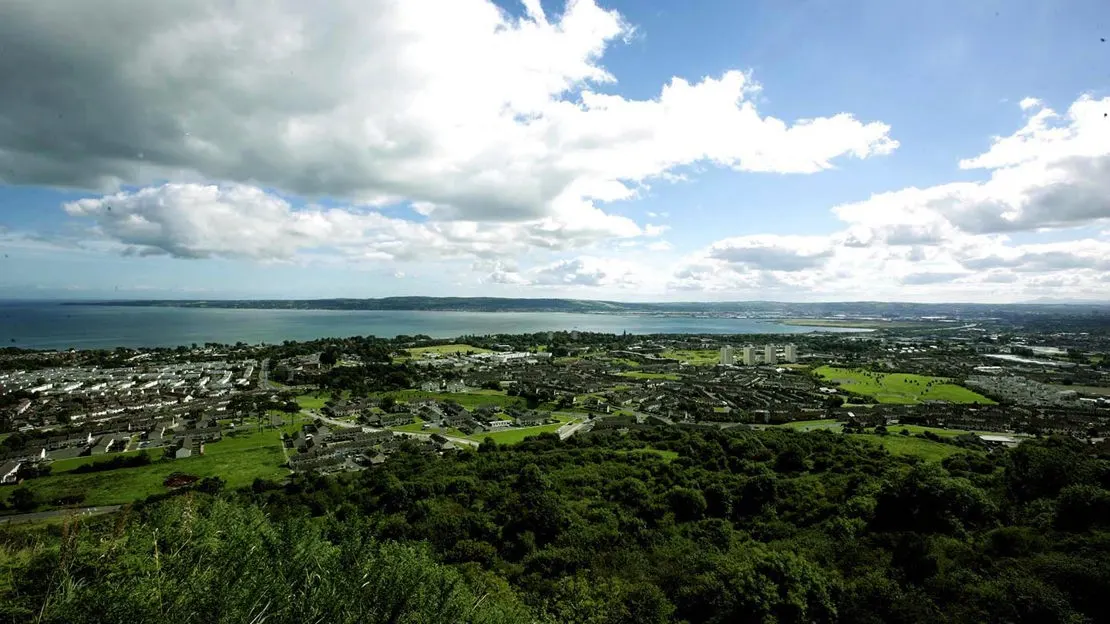 Aerial view from a hilltop overlooking a coastal town, green fields, and bay under blue sky with white clouds.