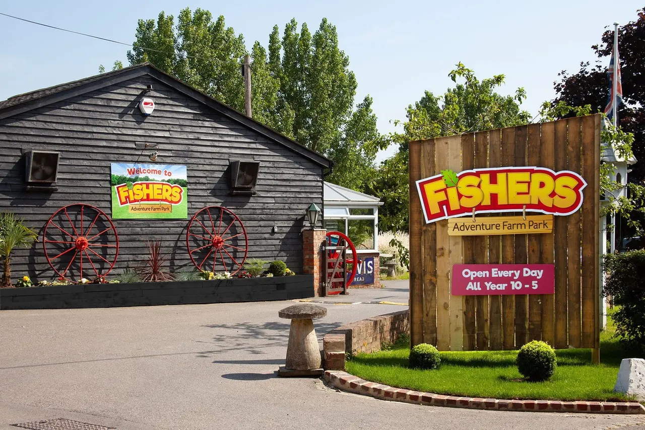 Entrance to Fishers Adventure Farm Park with signage, a barn building with red wagon wheels, and a wooden gate sign displaying opening hours.