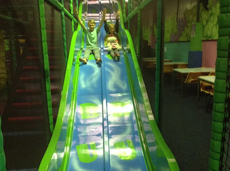 Two children sliding down a bright green and blue indoor slide at a play centre.