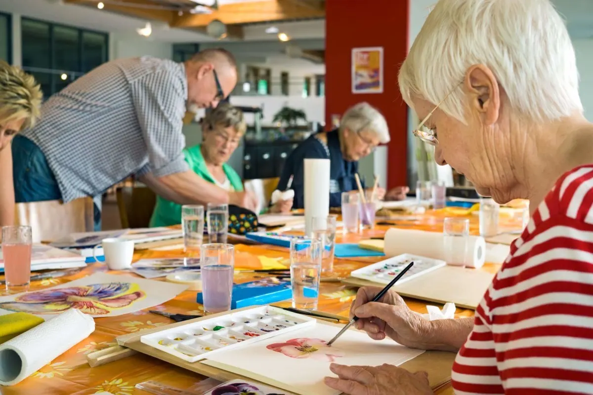 Older adults participate in a painting class at a table, with one woman in the foreground painting while others work nearby.