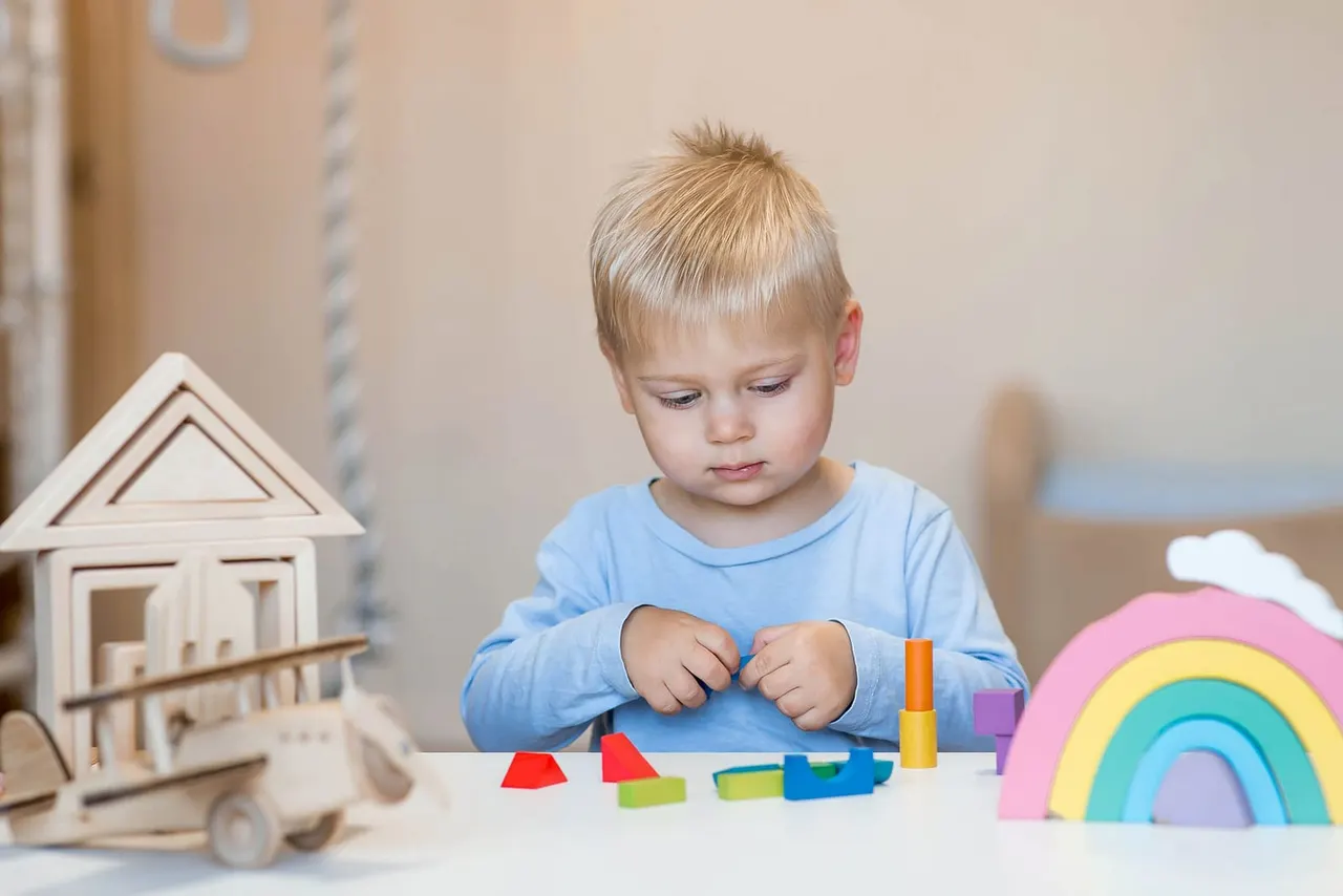 Young child in blue shirt playing with colourful wooden blocks and toys at a table.