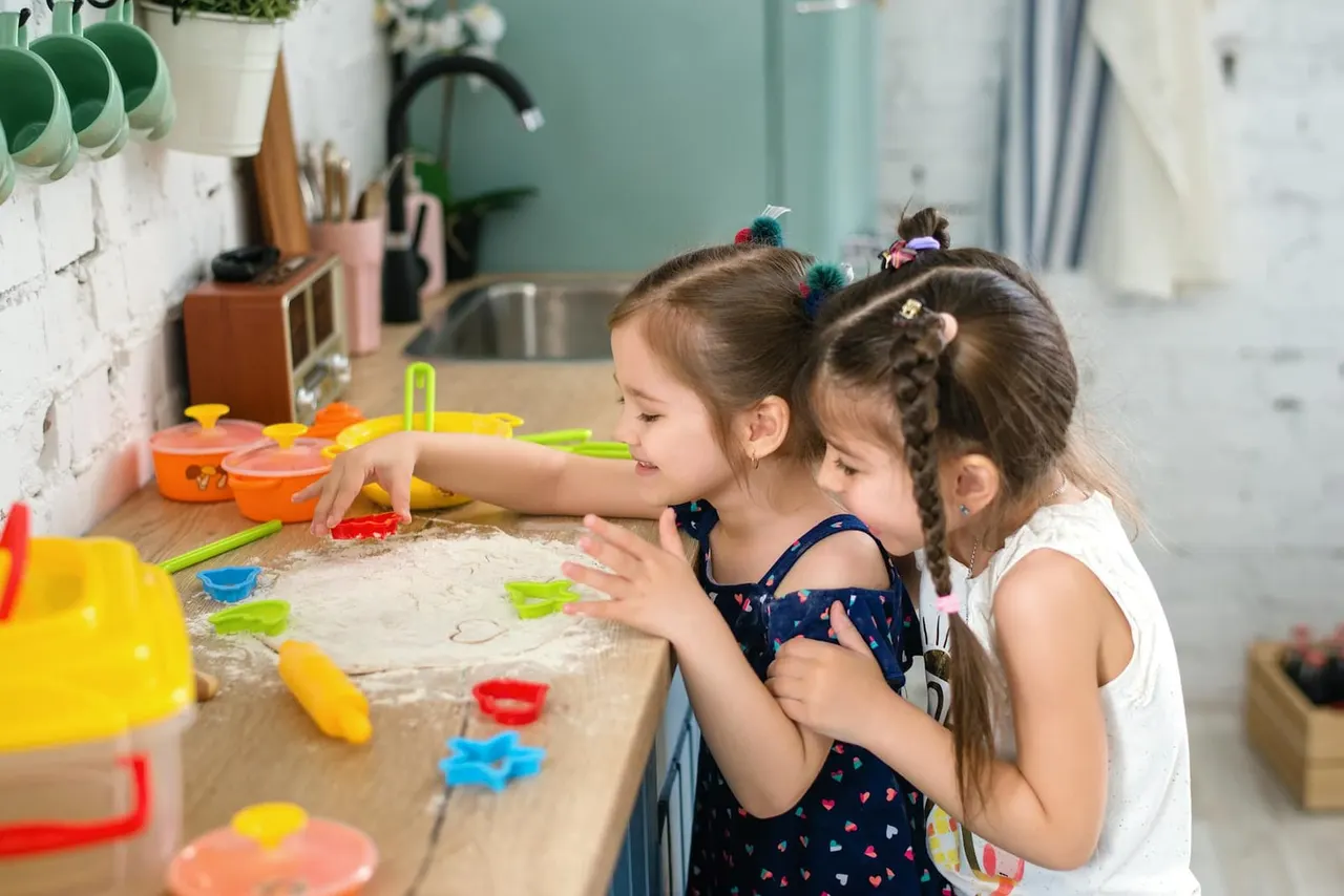 Two young children play in a sand table with colourful toy cookware and utensils in a bright kitchen-themed activity area.