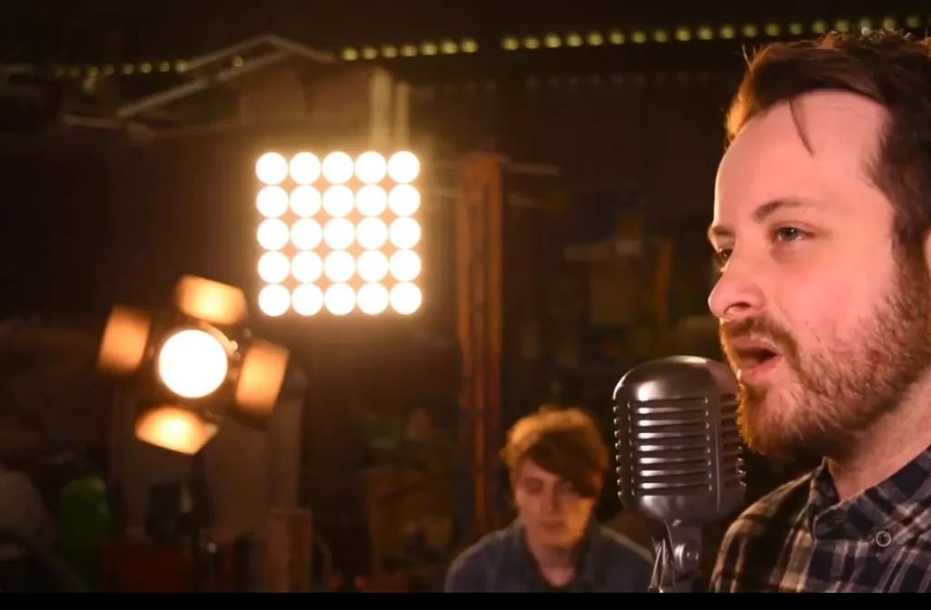 Man singing into vintage microphone on stage with theatrical lighting and audience members visible.