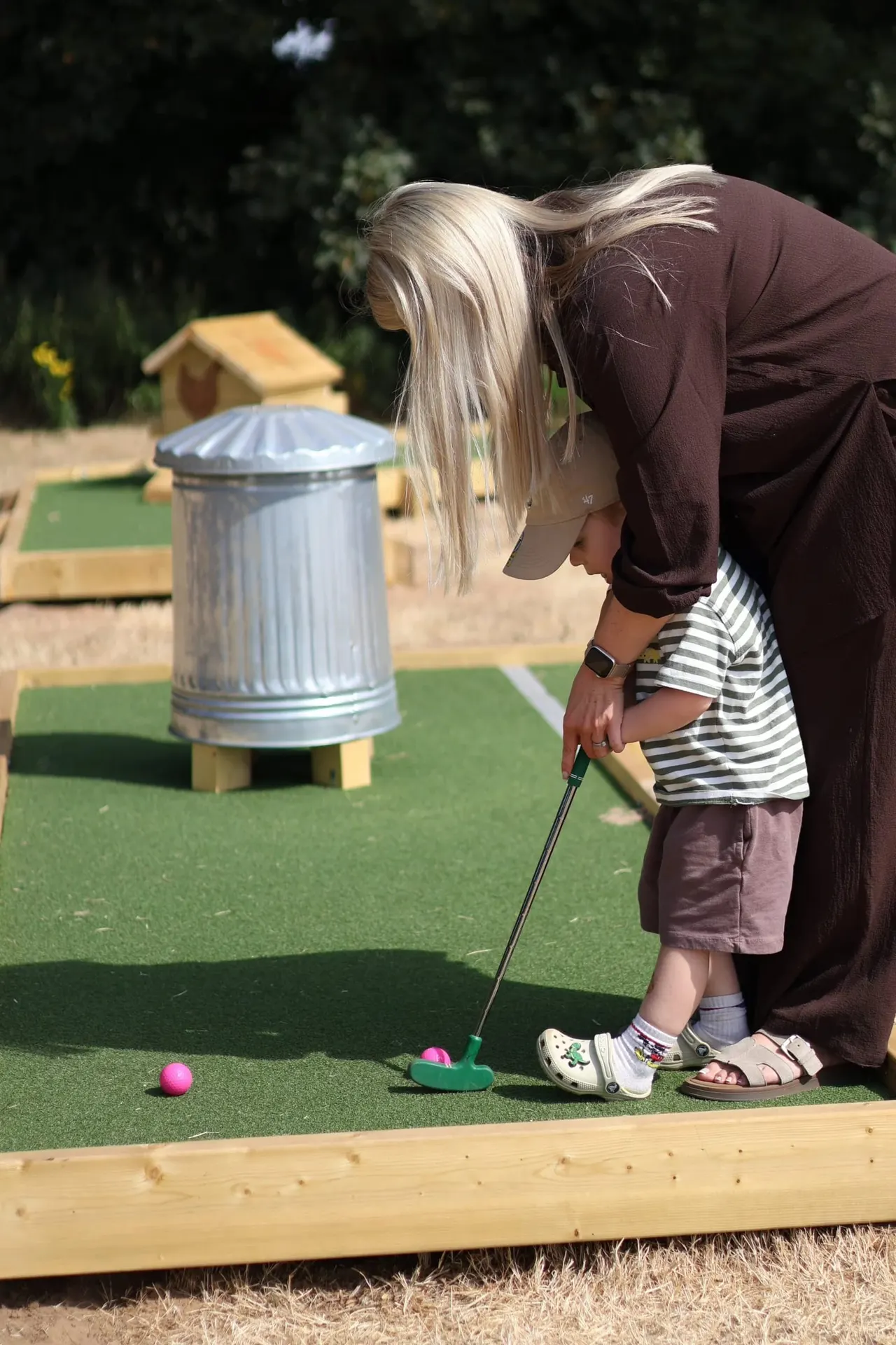 An adult helps a young child play mini golf on a bright green course with a pink ball and putter.