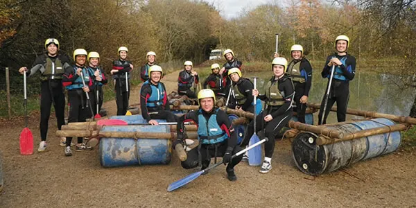 Large group of people wearing helmets and safety gear posing with paintball equipment and barrels outdoors.