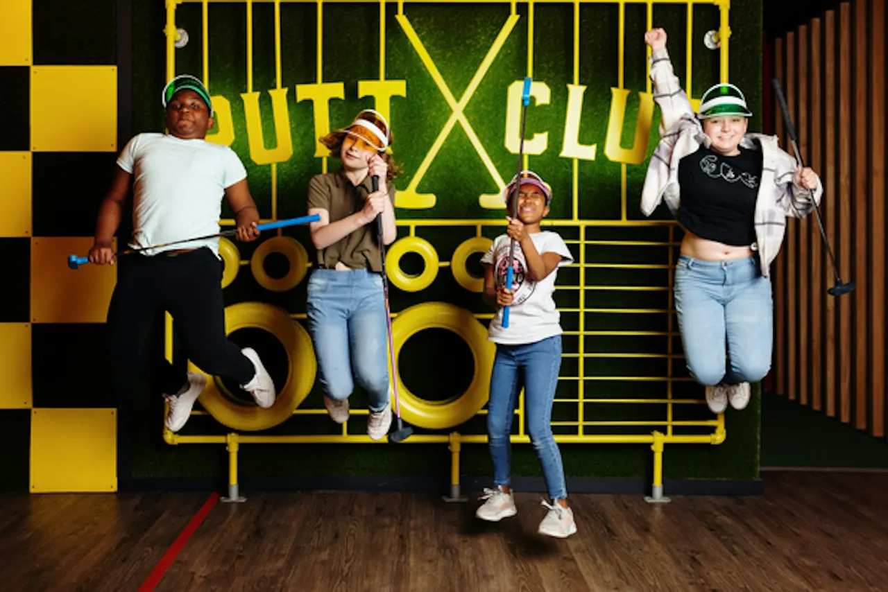 Four people of varying ages posing with baseball bats in front of a bright yellow and green neon-lit golf clubhouse backdrop.