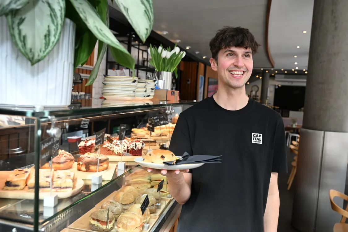 Smiling staff member holding a plate with a pastry in a modern cafe with display cases of baked goods and potted plants.