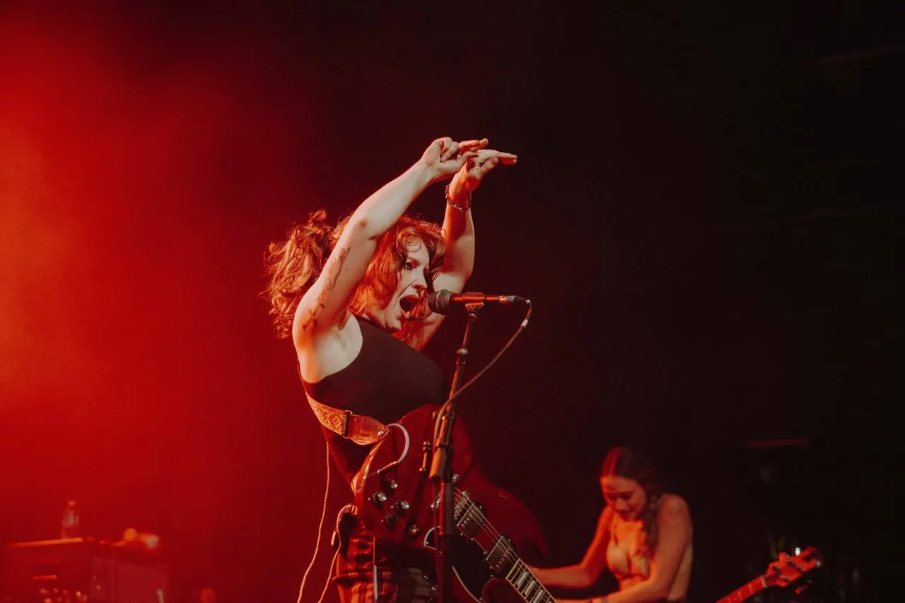 Female performer singing at microphone with raised arms during live music performance on stage lit in red.