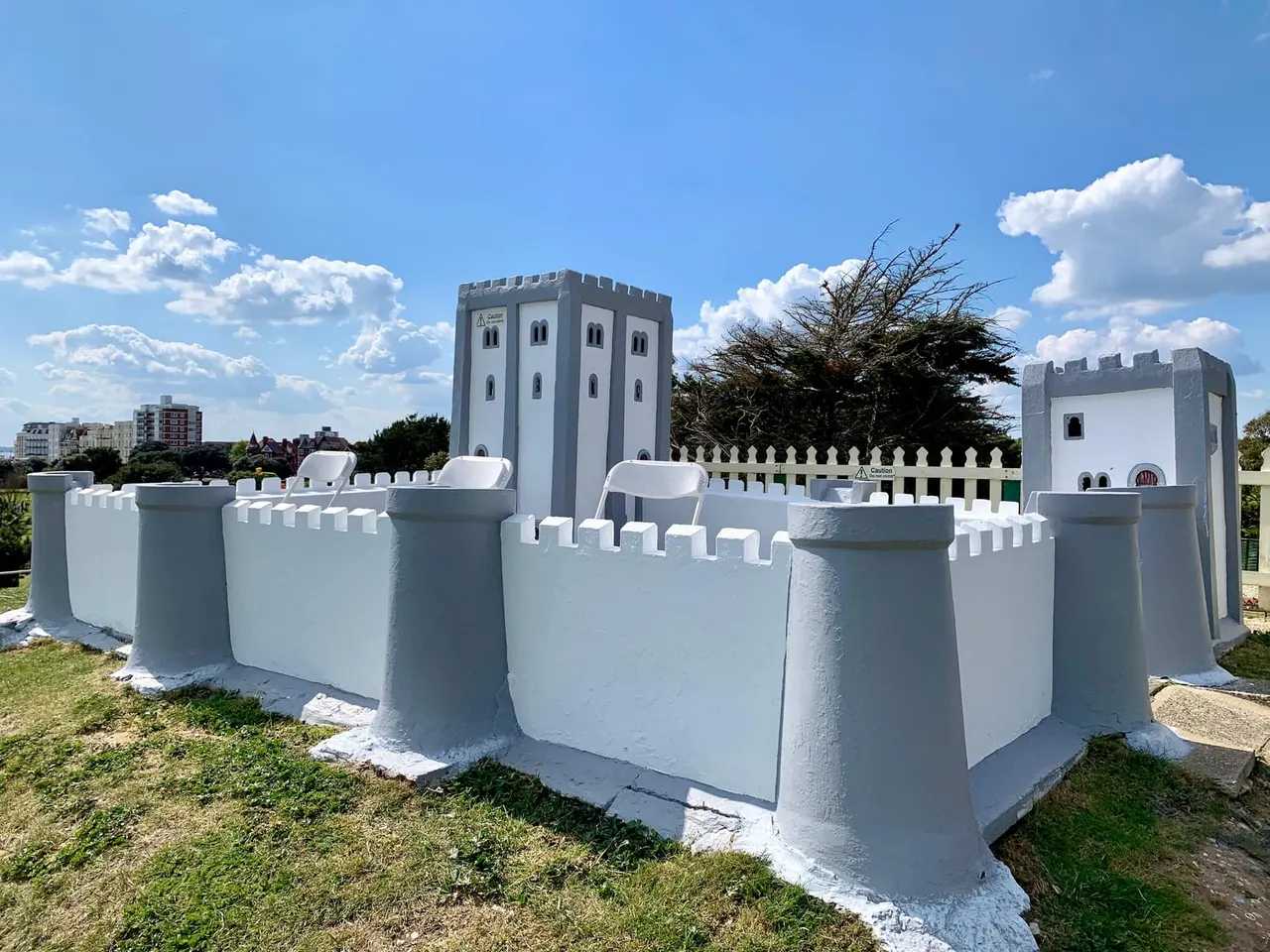 White-painted miniature castle fortification with crenellated walls and towers, set in a landscaped garden under blue sky.