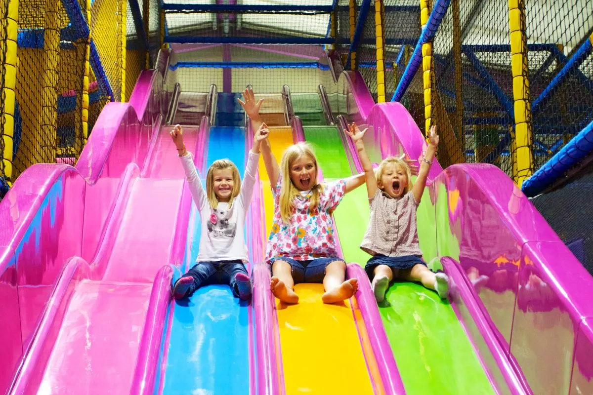 Three young girls sliding down brightly coloured chutes with arms raised, smiling excitedly in an indoor play centre.