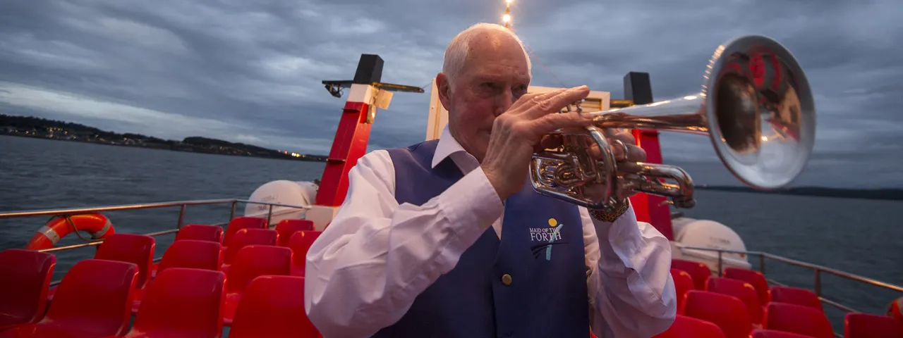 Man in formal waistcoat playing trumpet on the deck of a passenger ferry at dusk, with red seating visible behind him.