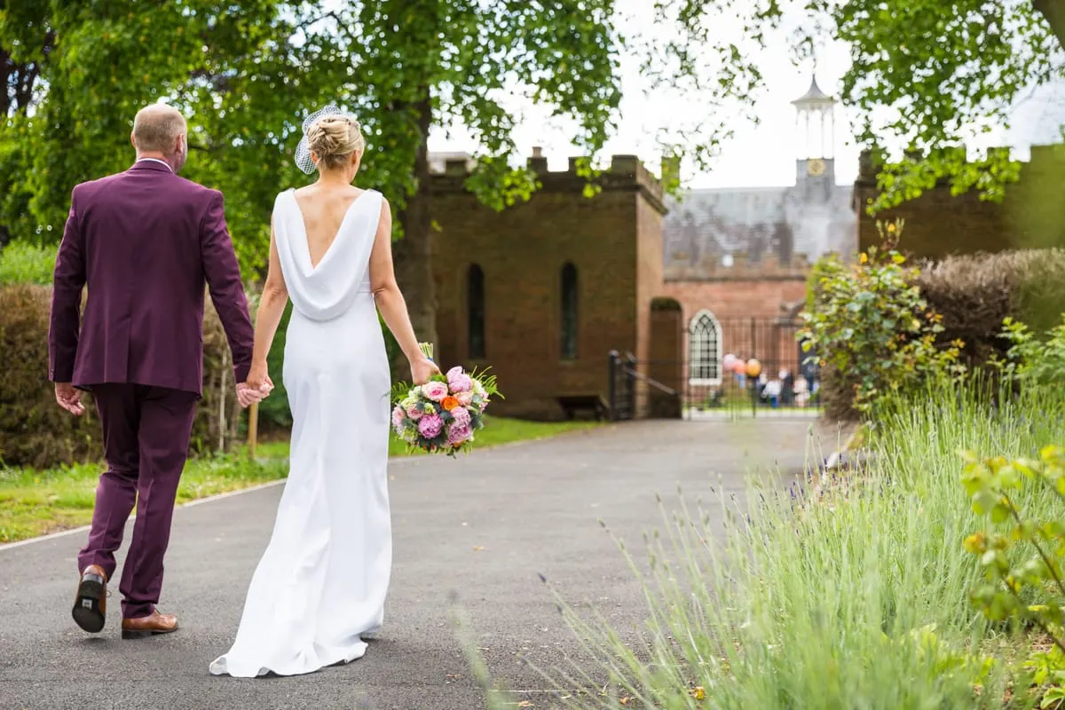 Bride and groom walk hand in hand along a garden path towards a castle, with the bride carrying a pink and orange bouquet.