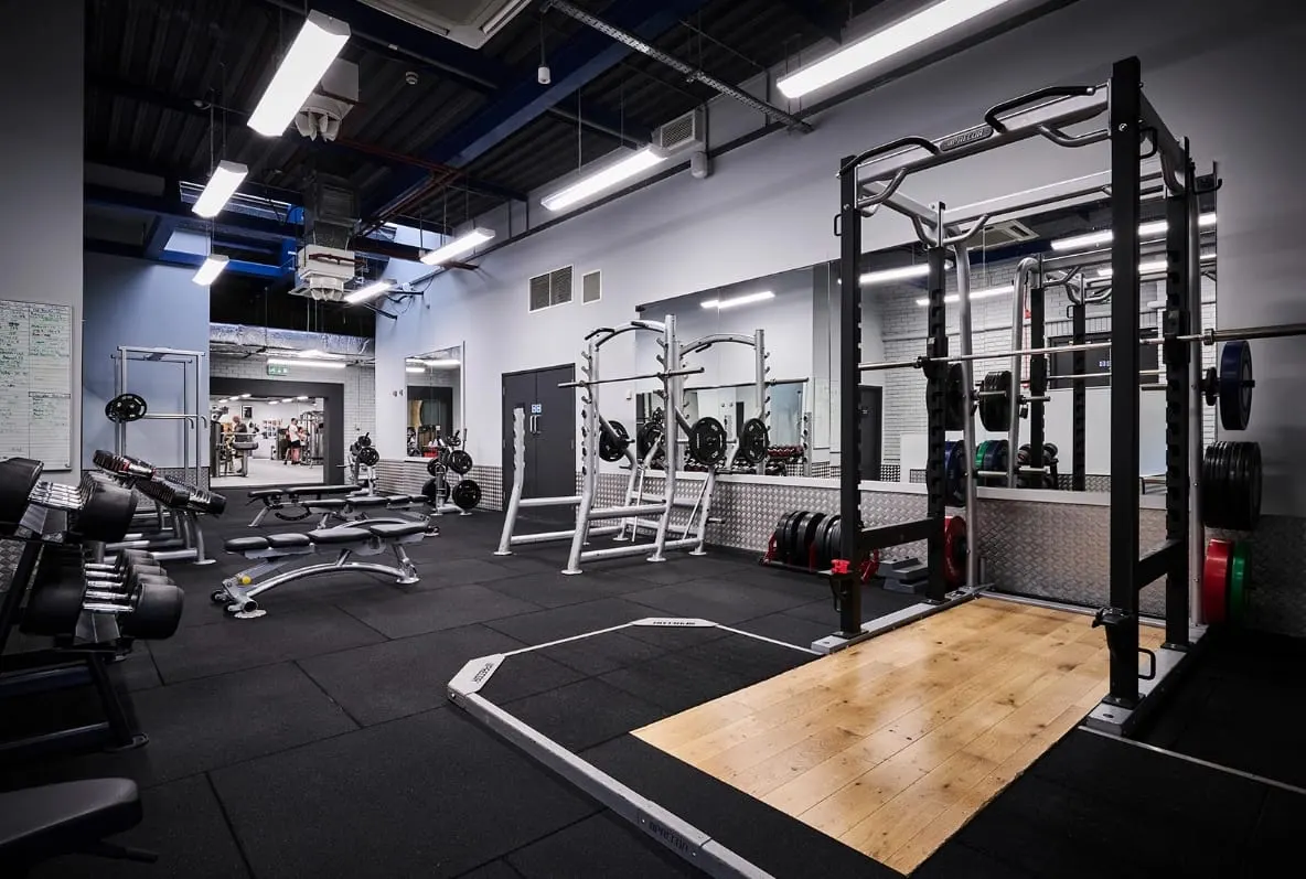 Modern gym interior with weight racks, dumbbells, exercise machines and wooden lifting platform on dark flooring.