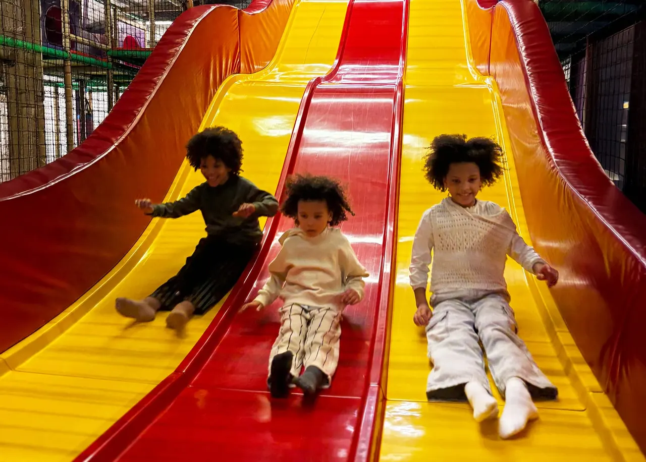 Three young children sliding down a colourful inflatable slide with red and yellow lanes indoors.