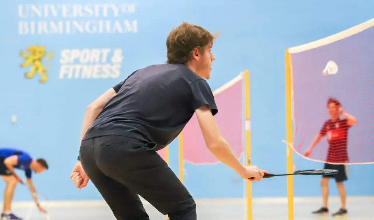 Young person playing badminton indoors with opponents visible through the net.