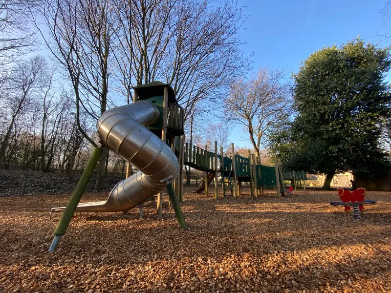 Spiral metal slide and playground equipment in a woodland play area with bare trees and clear blue sky.