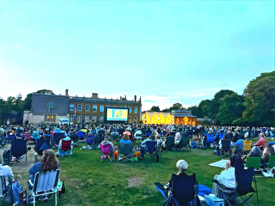 Large outdoor crowd seated on grass watching a film on a giant screen projected on a historic building's facade.