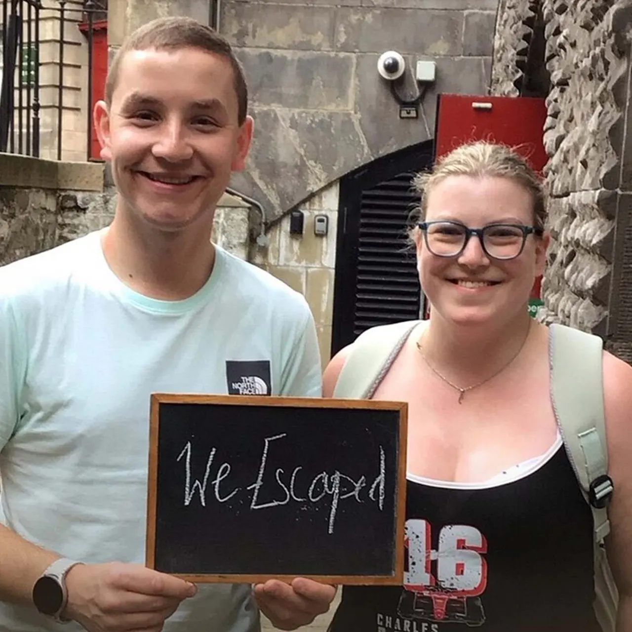 Two young people smile at the camera whilst holding a small blackboard reading "We Escaped" outside a stone building.