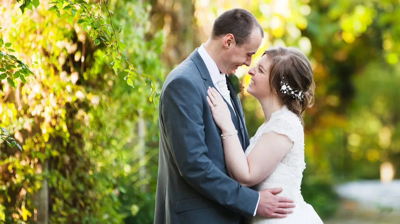 Bride and groom smiling at each other among ivy-covered walls in a sunlit garden setting.