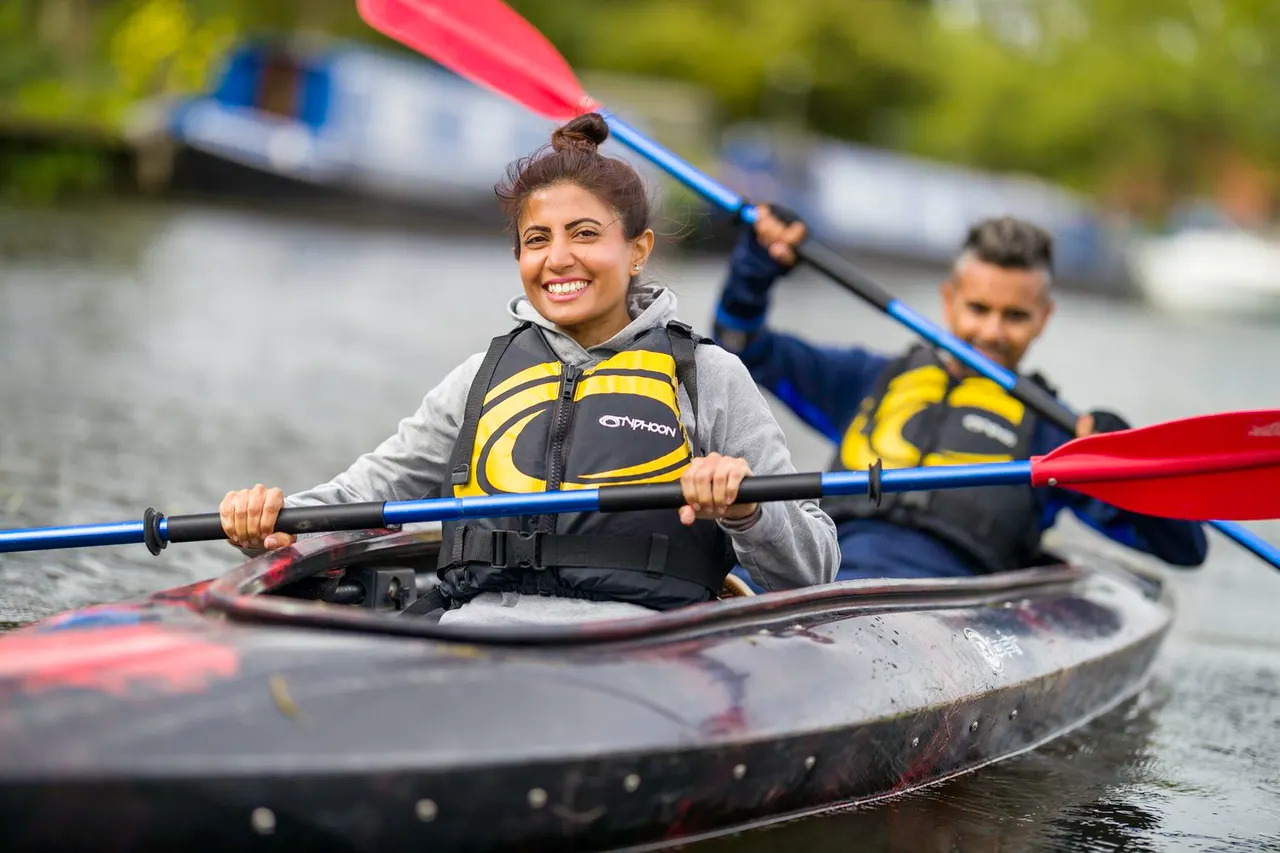 Woman and young person kayaking on a river, wearing yellow and black life jackets and holding paddles.