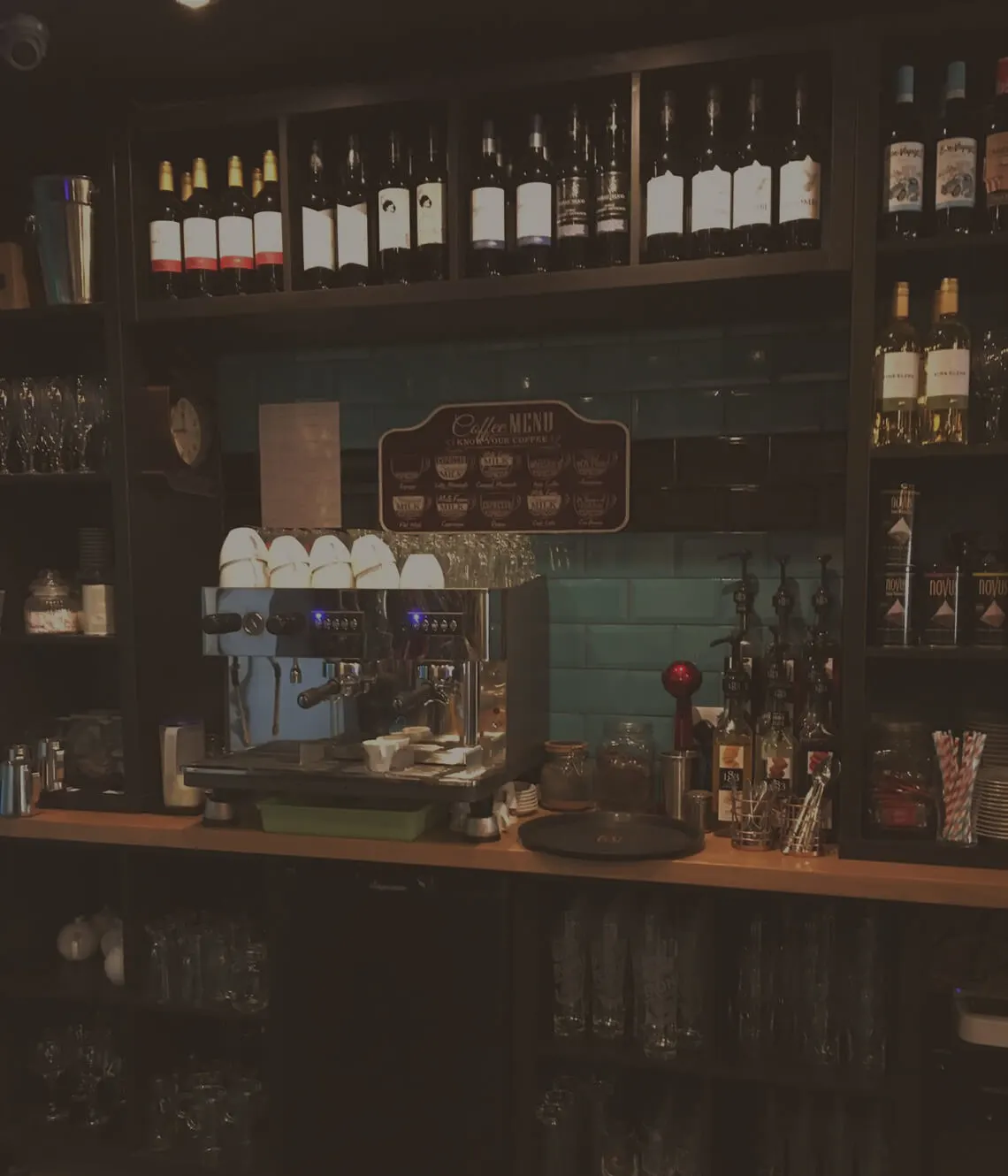 A cafe bar counter with an espresso machine, coffee menu board, and shelves of wine bottles and spirits displayed behind.