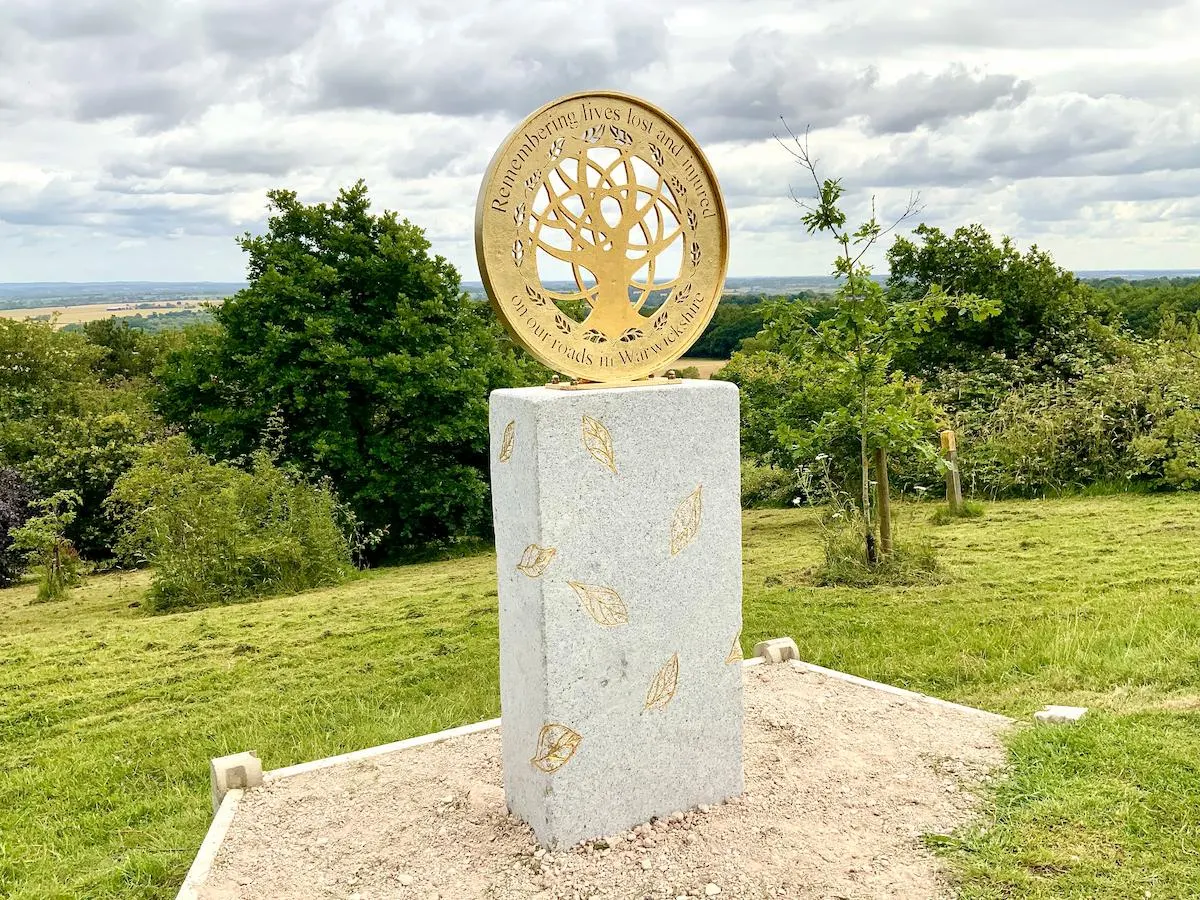 Gold tree sculpture on stone plinth overlooking forested countryside landscape under cloudy sky.