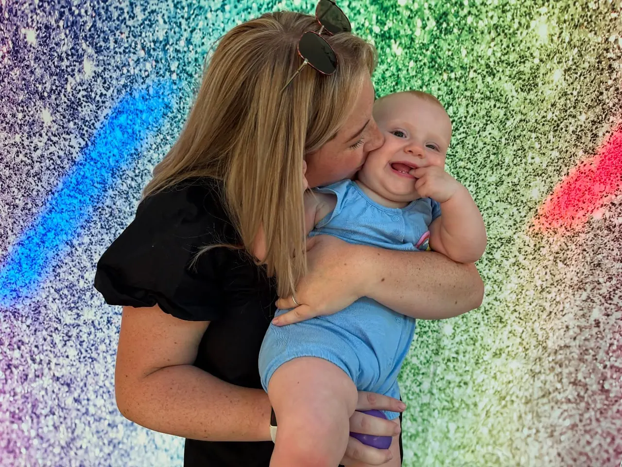 Woman holding a smiling baby in front of a colourful sequined backdrop.