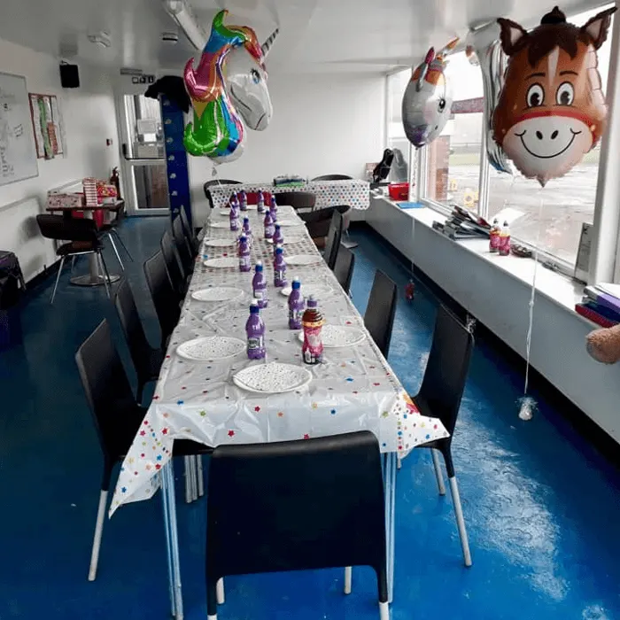 A party room decorated with colourful balloons and a long table set with plates and drinks for children's celebration.