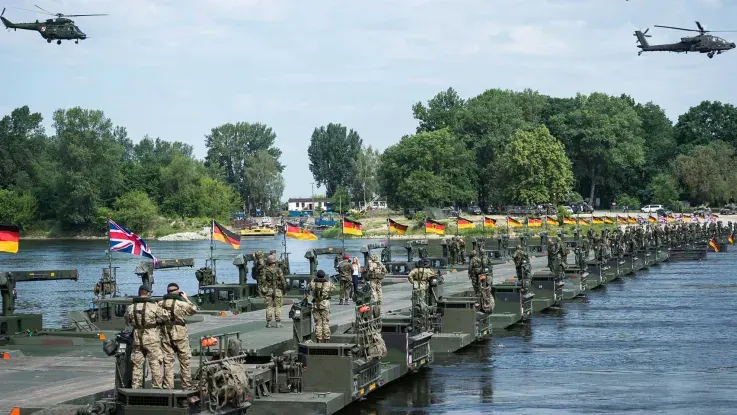 Military personnel and pontoon bridge sections assembled in water with flags and helicopters overhead.