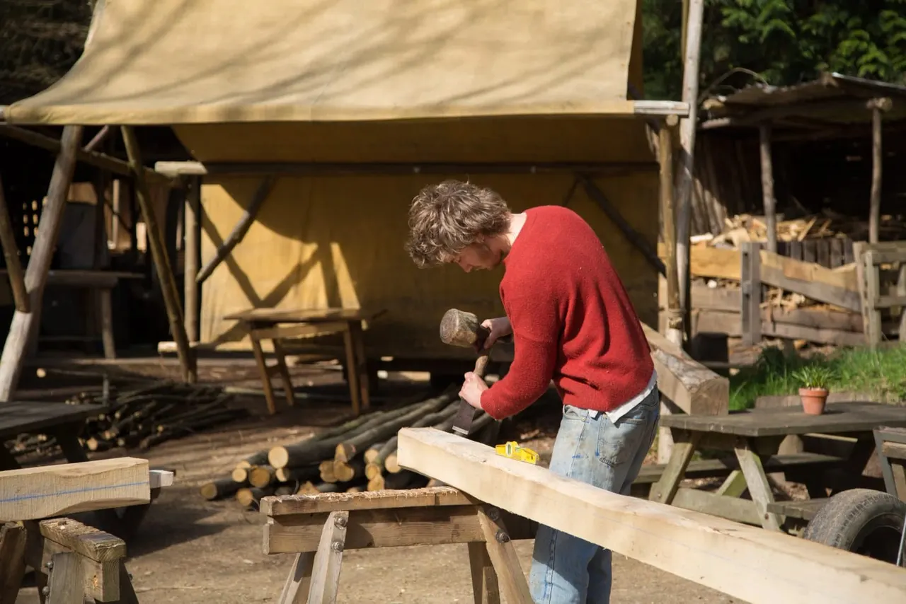 A person in a red jumper chiselling wood at an outdoor workshop beneath canvas awnings.
