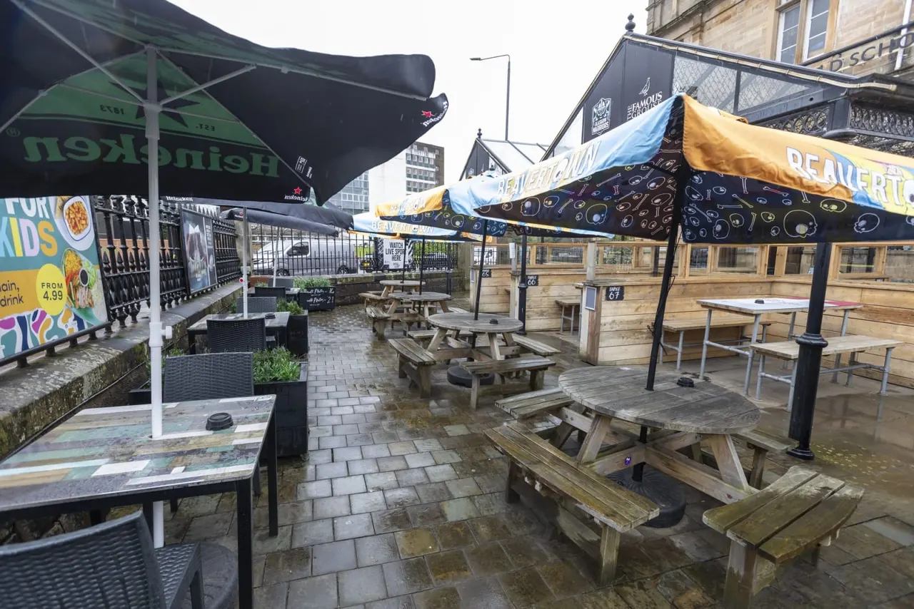 Outdoor terrace with picnic tables and umbrellas in a wet urban setting surrounded by stone buildings.