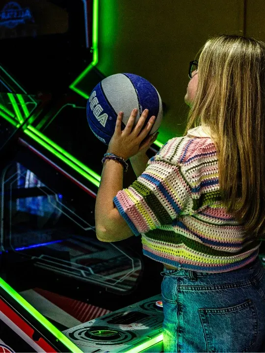 Young person holding a basketball at a neon-lit arcade game with green and blue lighting.