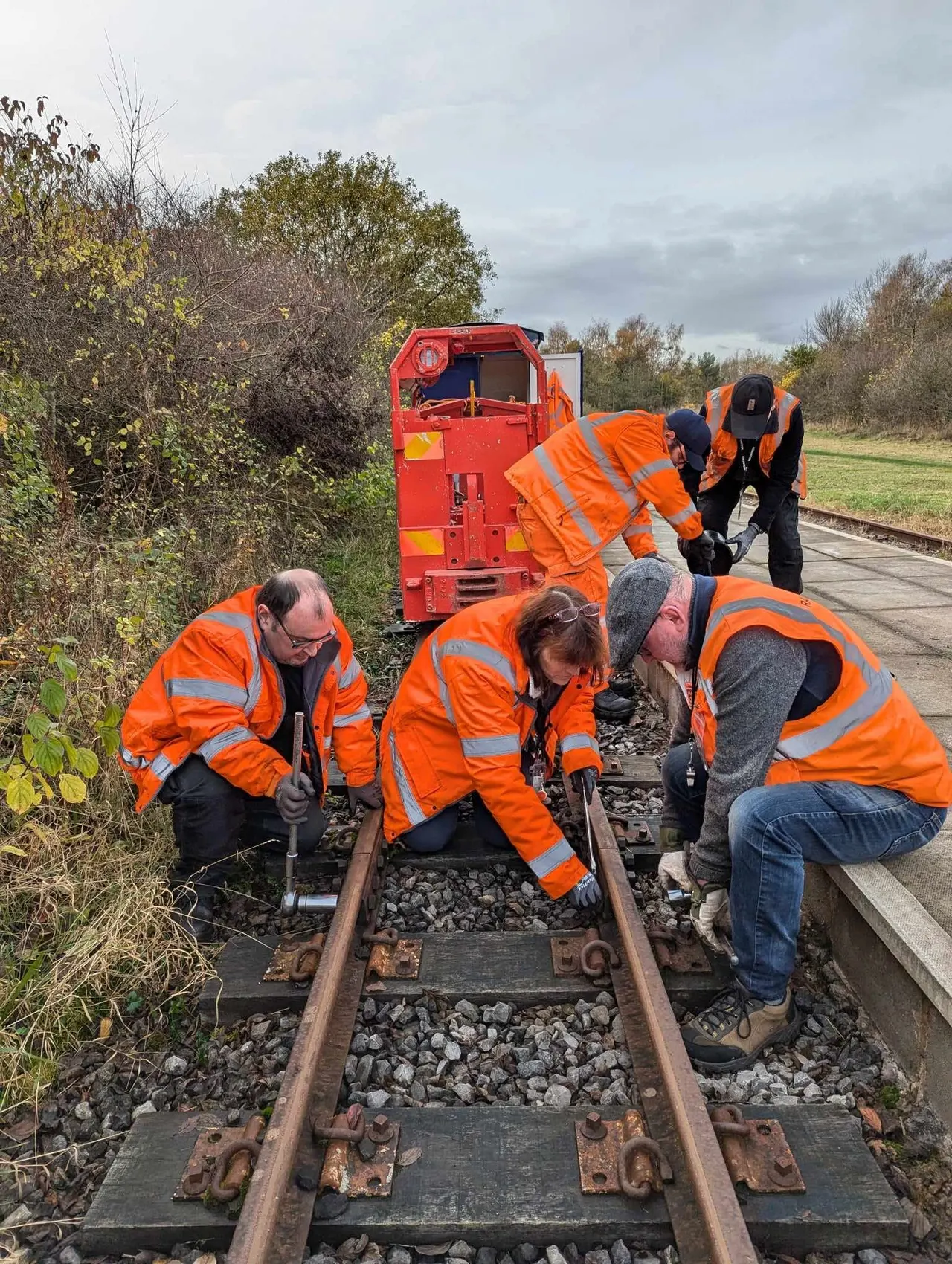 Workers in orange high-visibility jackets maintain railway tracks alongside a red maintenance vehicle in an autumn landscape.