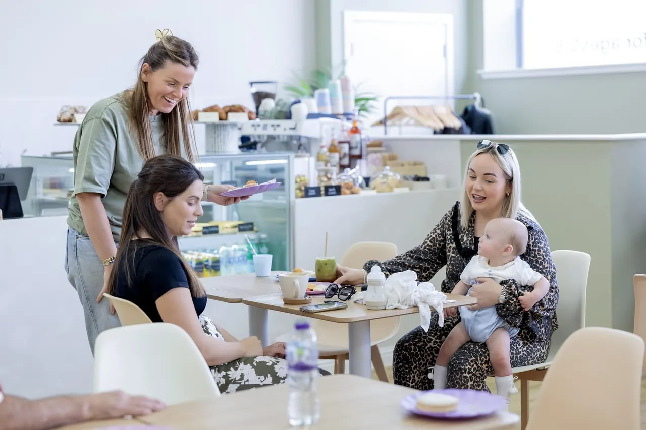 Staff member serving food to a woman with a baby and another woman at a table in a bright café.