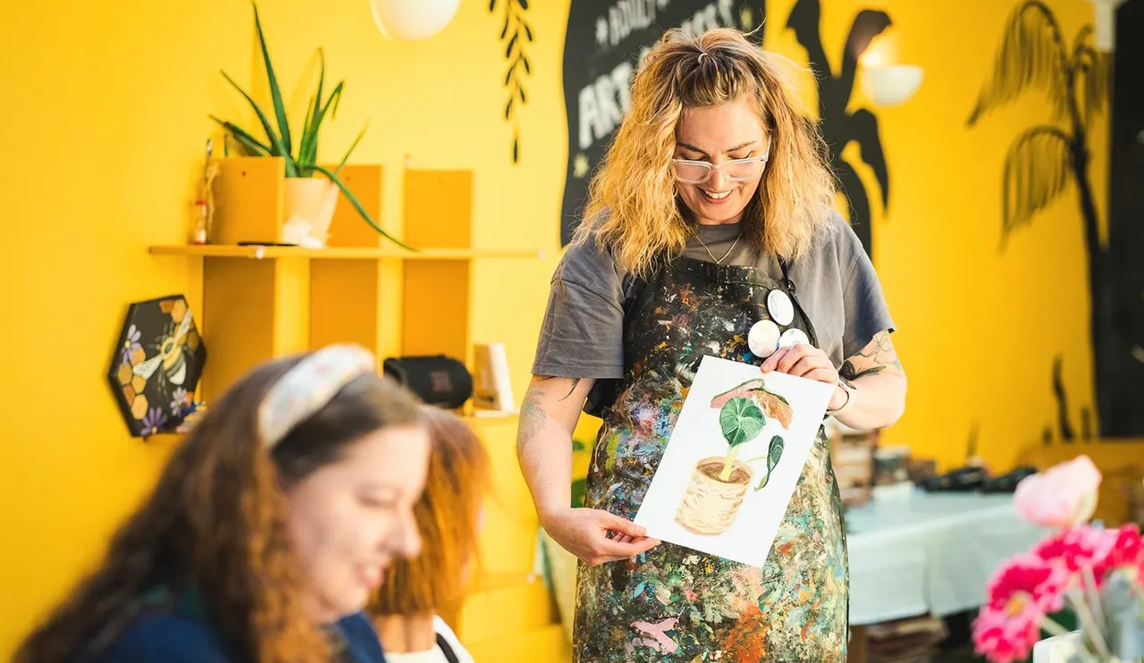 Woman showing artwork to visitor in bright yellow studio with potted plants and botanical decorations.