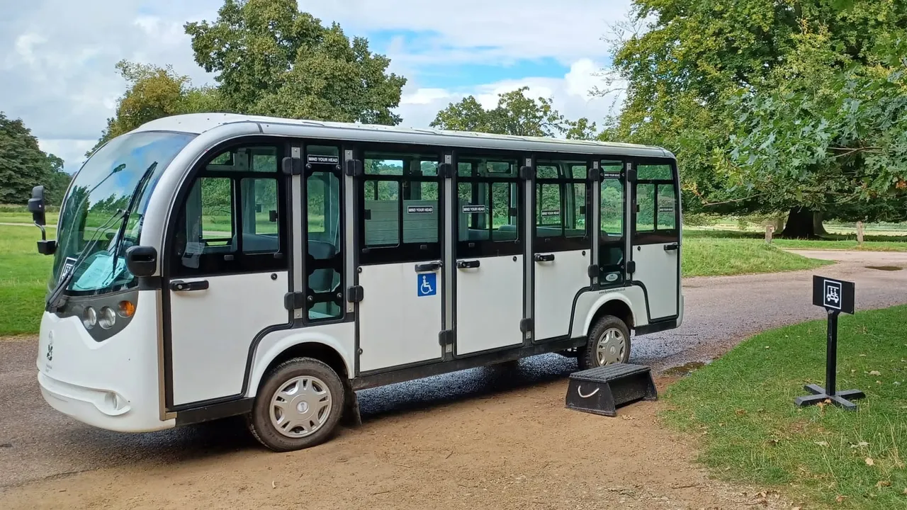 White and black accessible shuttle bus parked on a tree-lined path in parkland, with disability access ramp deployed.
