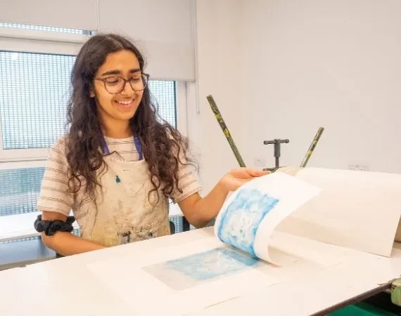 A young person wearing glasses and a paint-splattered apron demonstrates a printing or art technique using brushes and paper at a worktable.