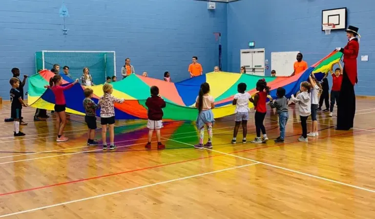 Children and adults playing with a large colourful parachute in a sports hall during a group activity session.