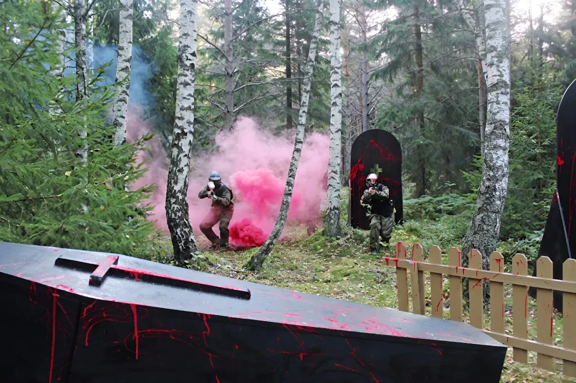 Players in camouflage gear surrounded by pink powder smoke during a paintball match in a forest setting.