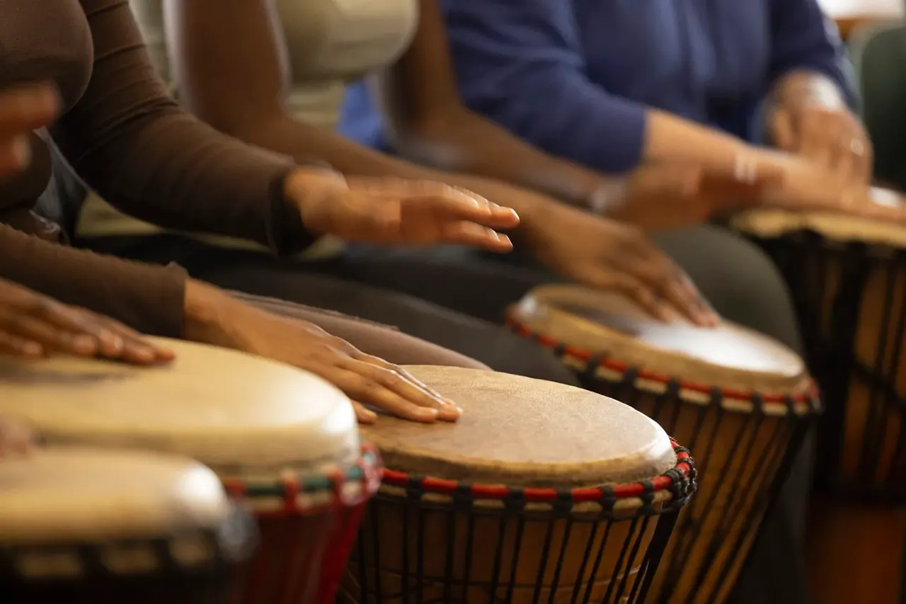 Multiple people's hands playing frame drums with red and natural-coloured rope binding during a workshop or performance.