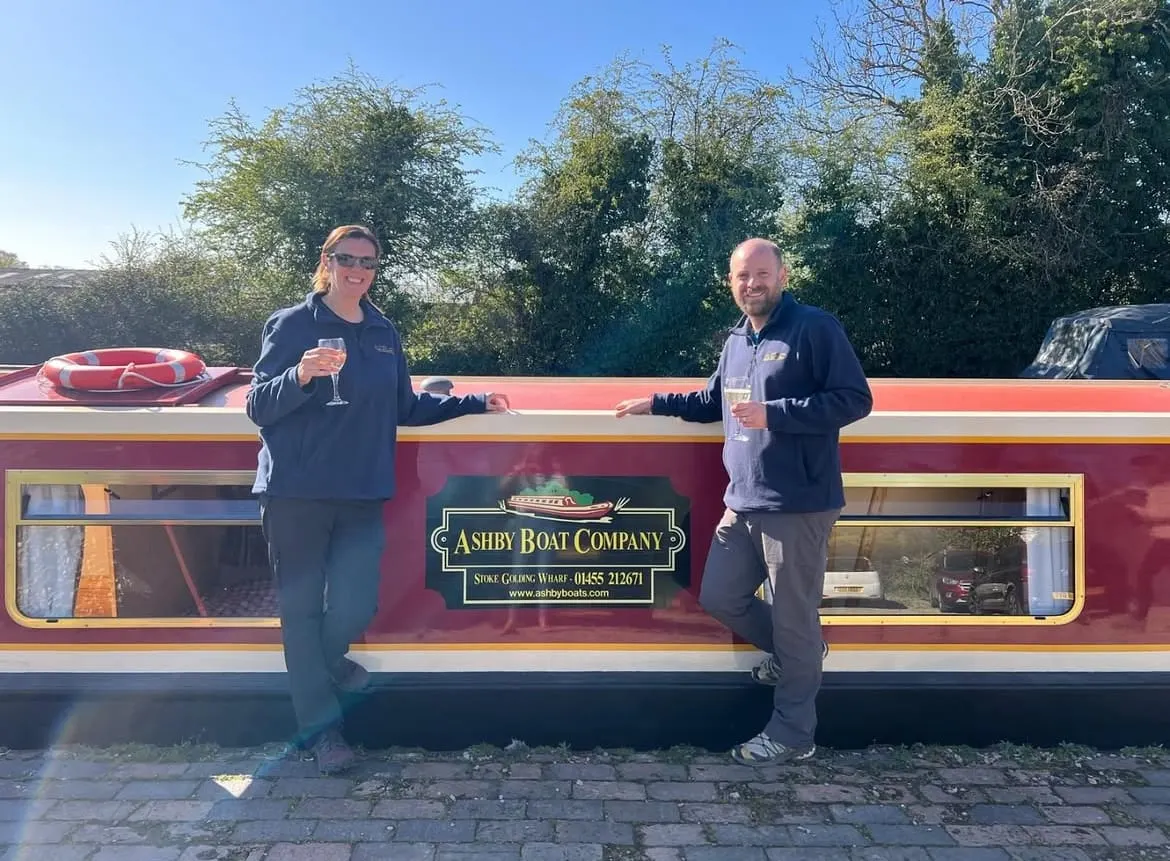 Two people holding drinks stand outside a red and gold narrowboat moored on a canal, with trees in the background.