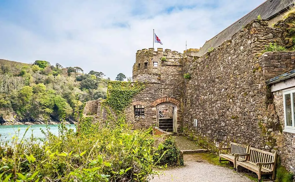 Stone castle with Union flag flying from central tower, overlooking river estuary with wooded hillside, wooden benches in foreground.