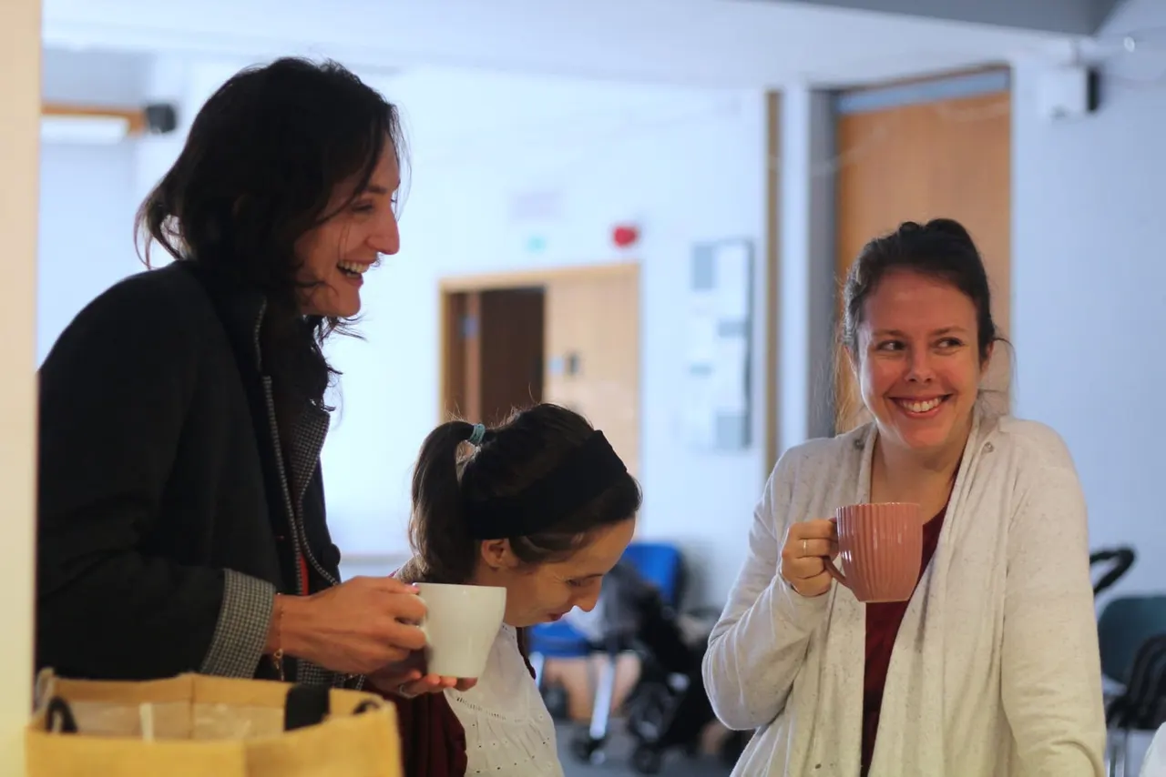 Two women and a child holding cups enjoy refreshments together in a bright community hall.
