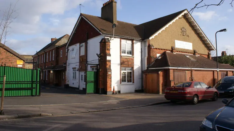 Exterior of a brick building with white sections and green gates, parked cars in front on a residential street.