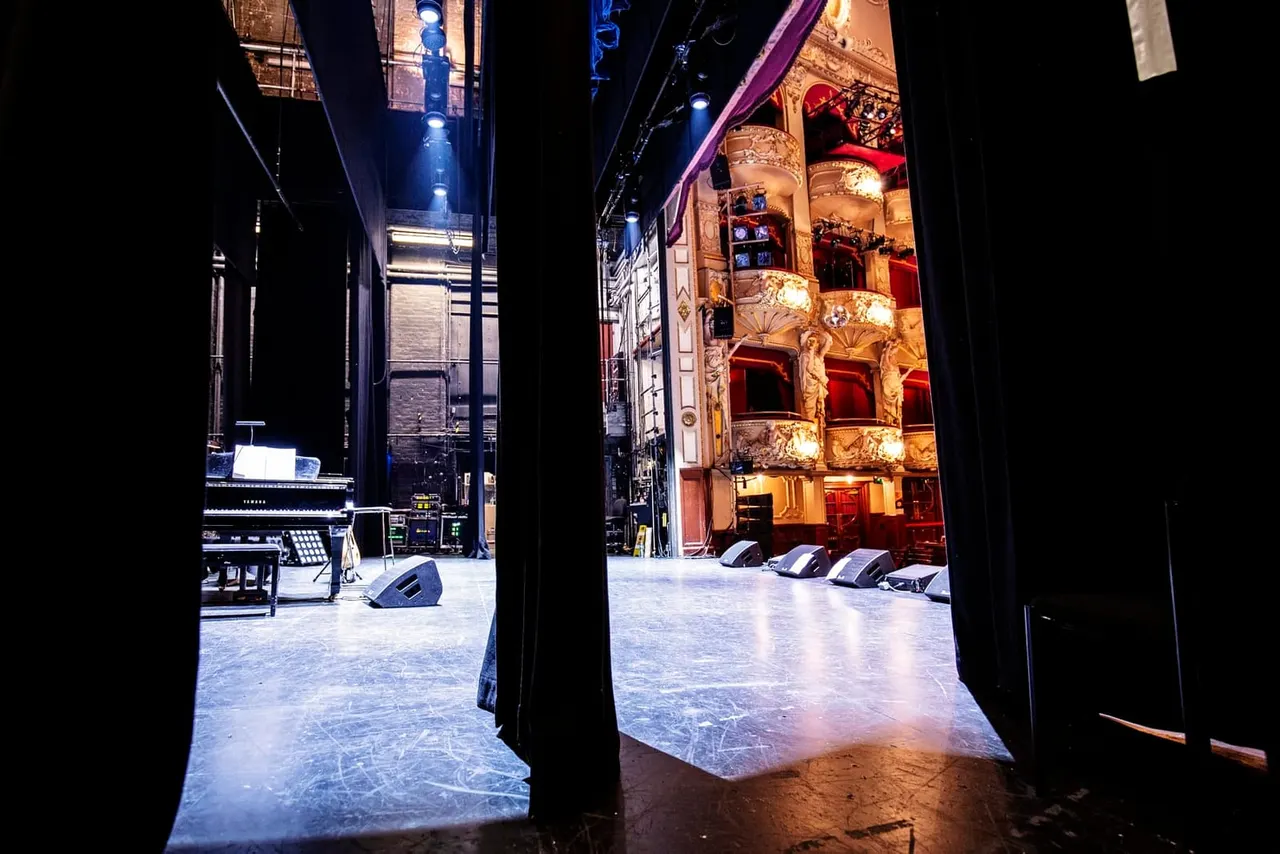 Theatre stage with ornate gilt boxes and red seating visible, lit by warm and blue stage lighting at night.