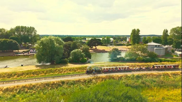 A narrow-gauge passenger train travels through open countryside beside a river lined with trees and farm buildings.