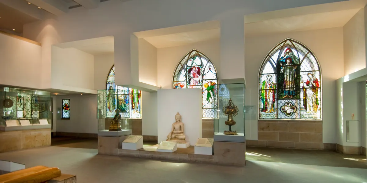 Museum gallery with stained glass windows and display cases containing sculptures and religious artefacts.