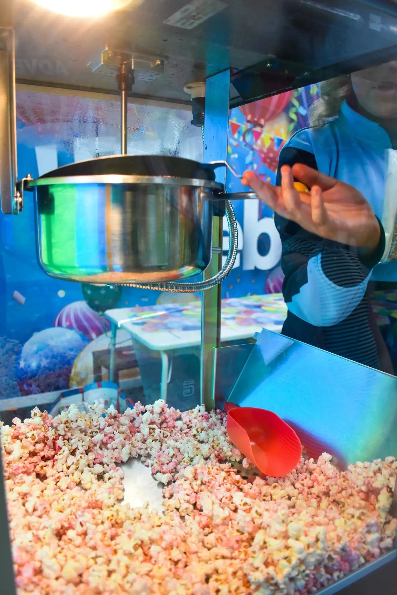 Person in blue shirt operating a popcorn machine with coloured popcorn spilling out below.